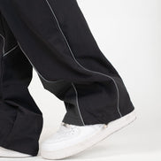 Close-up of a person's feet wearing black wide-leg pants with contrasting light gray piping and white low-top sneakers against a clean white studio background.