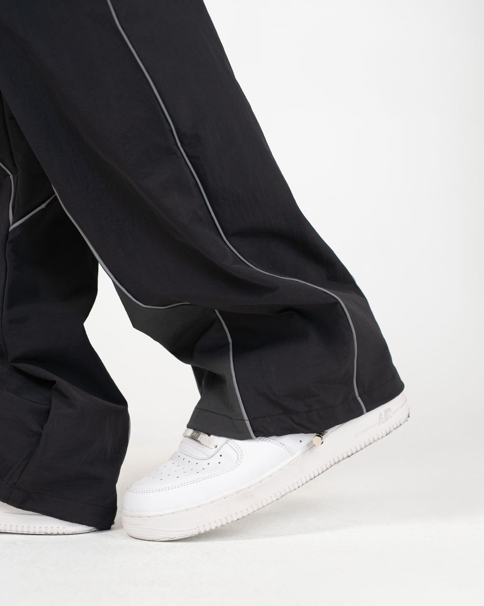 Close-up of a person's feet wearing black wide-leg pants with contrasting light gray piping and white low-top sneakers against a clean white studio background.