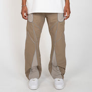 A person wearing loose-fit beige cargo-style pants with contrasting light gray paneling and white sneakers, standing against a plain light gray studio background.
