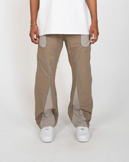 A person wearing loose-fit beige cargo-style pants with contrasting light gray paneling and white sneakers, standing against a plain light gray studio background.