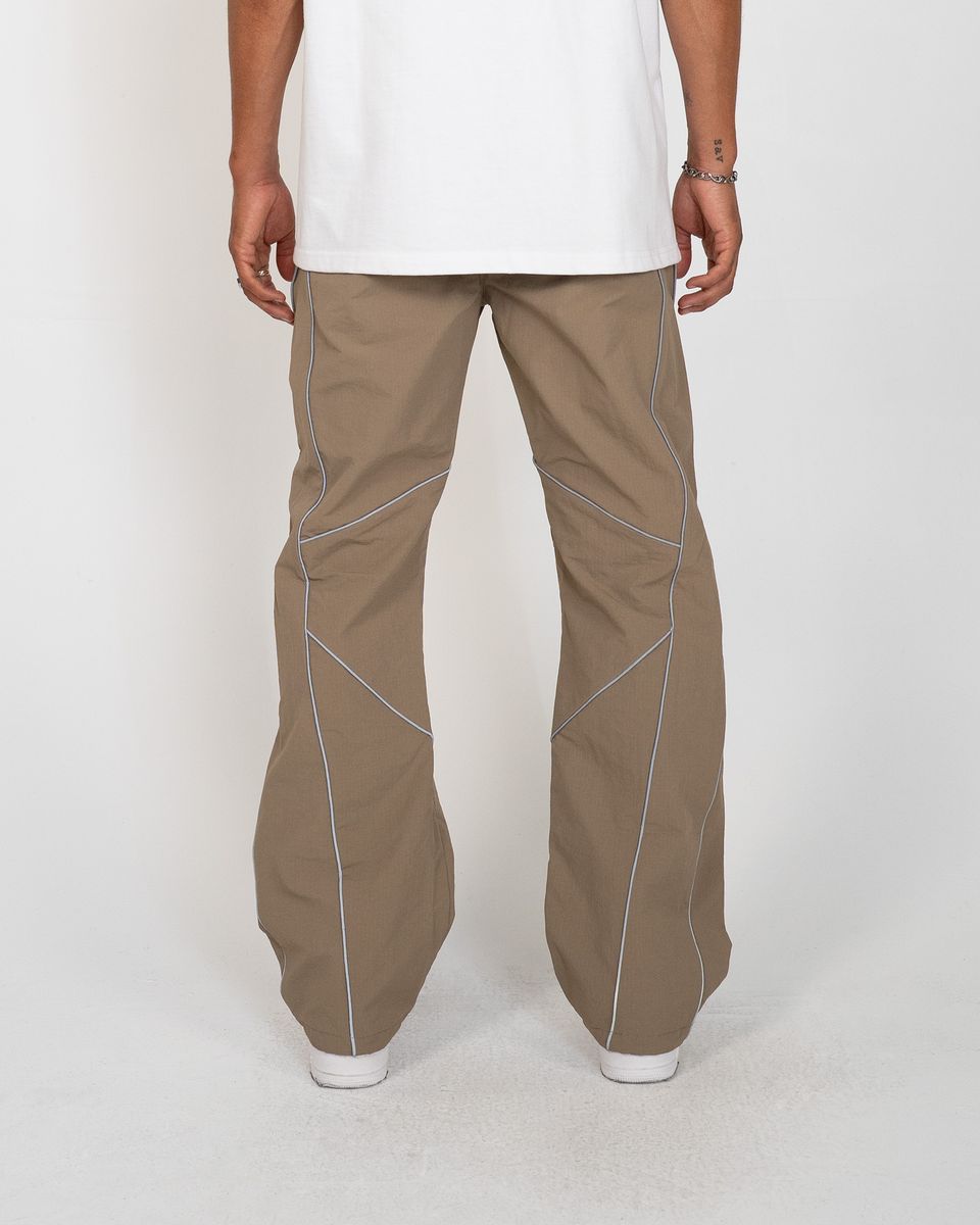A person shown from the back wearing loose-fit taupe-brown pants with contrasting light blue piping details, a white t-shirt, and white sneakers against a light gray studio background.