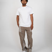 A man modeling a white short-sleeve crewneck t-shirt and light brown pants with contrasting light gray side panels, paired with white sneakers, against a plain light gray studio background.