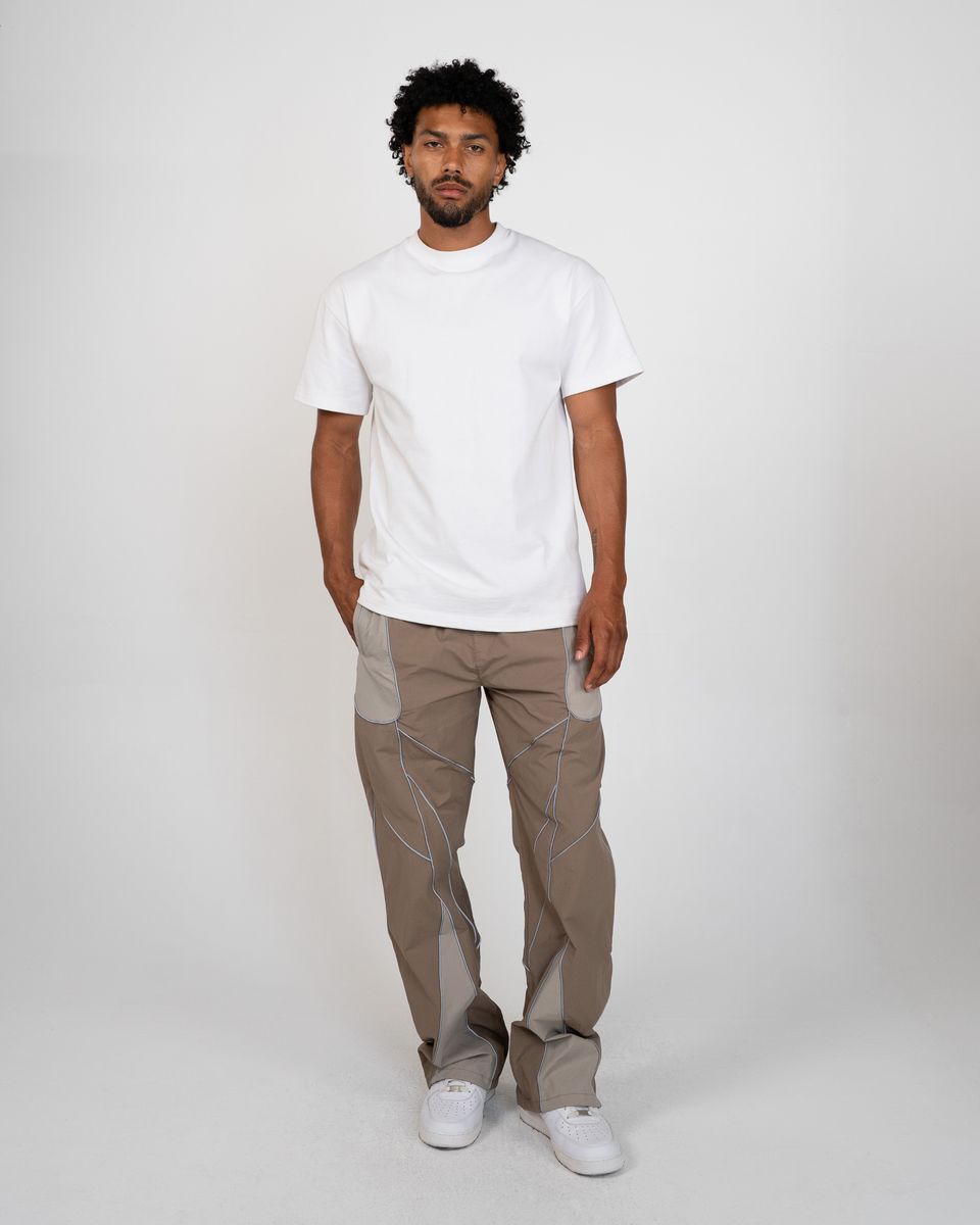 A man modeling a white short-sleeve crewneck t-shirt and light brown pants with contrasting light gray side panels, paired with white sneakers, against a plain light gray studio background.