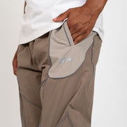 Close-up of a person wearing taupe-brown technical outdoor pants with contrast stitching and a white t-shirt, showing a side pocket with a light gray lining and a small embroidered logo.
