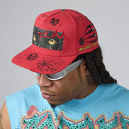 A red baseball cap with black graffiti-style drawings, a graphic eye patch, and yellow text reading 'PROTECT YOUR' worn by a person in a light blue graphic t-shirt against a gray studio background.