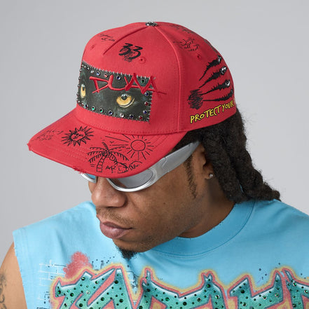 A red baseball cap with black graffiti-style drawings, a graphic eye patch, and yellow text reading 'PROTECT YOUR' worn by a person in a light blue graphic t-shirt against a gray studio background.