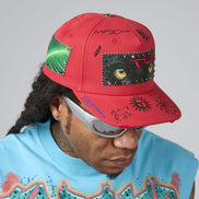 A red baseball cap with a green leaf patch, a black cat-face patch with silver studs, and various black script and graphic prints, worn by a model in a light blue graphic t-shirt against a gray background.