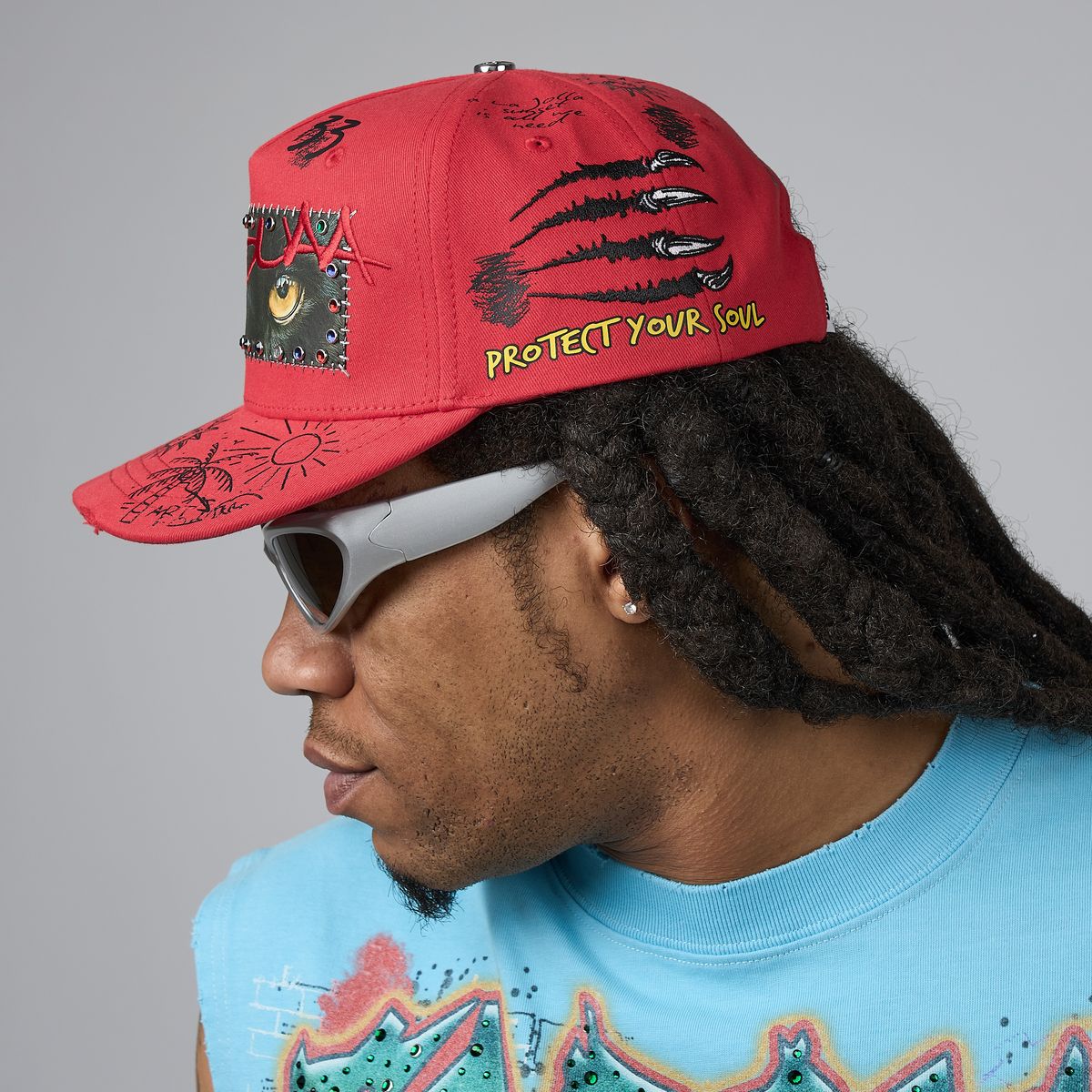A man in profile wearing a red baseball cap with black graphic designs, a patch on the side, and yellow embroidered text reading 'PROTECT YOUR SOUL'. He is also wearing white sunglasses and a light blue graphic t-shirt against a plain gray background.