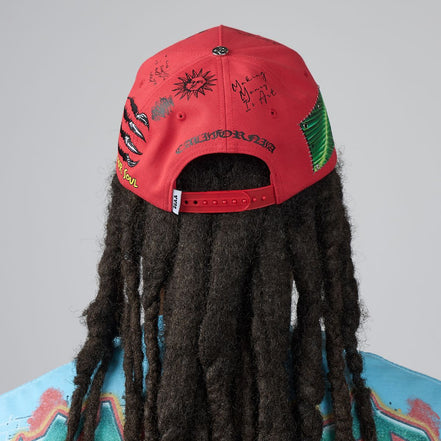 A red baseball cap with graffiti-style drawings and the word 'CALIFORNIA' on the back, shown from behind on a model with long dark dreadlocks against a light gray background.