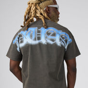 A man with blonde dreadlocks and white sunglasses, viewed from behind, wearing a dark gray t-shirt with a large light blue and white graphic print on the back, paired with pink patterned shorts, against a light gray background.
