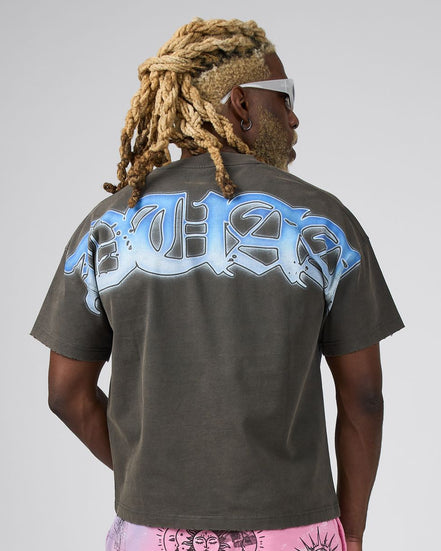 A man with blonde dreadlocks and white sunglasses, viewed from behind, wearing a dark gray t-shirt with a large light blue and white graphic print on the back, paired with pink patterned shorts, against a light gray background.