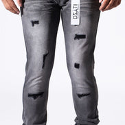 A person wearing distressed gray slim-fit jeans with ripped details and a white vertical brand tag on the thigh, paired with a white t-shirt and white sneakers against a white studio background.