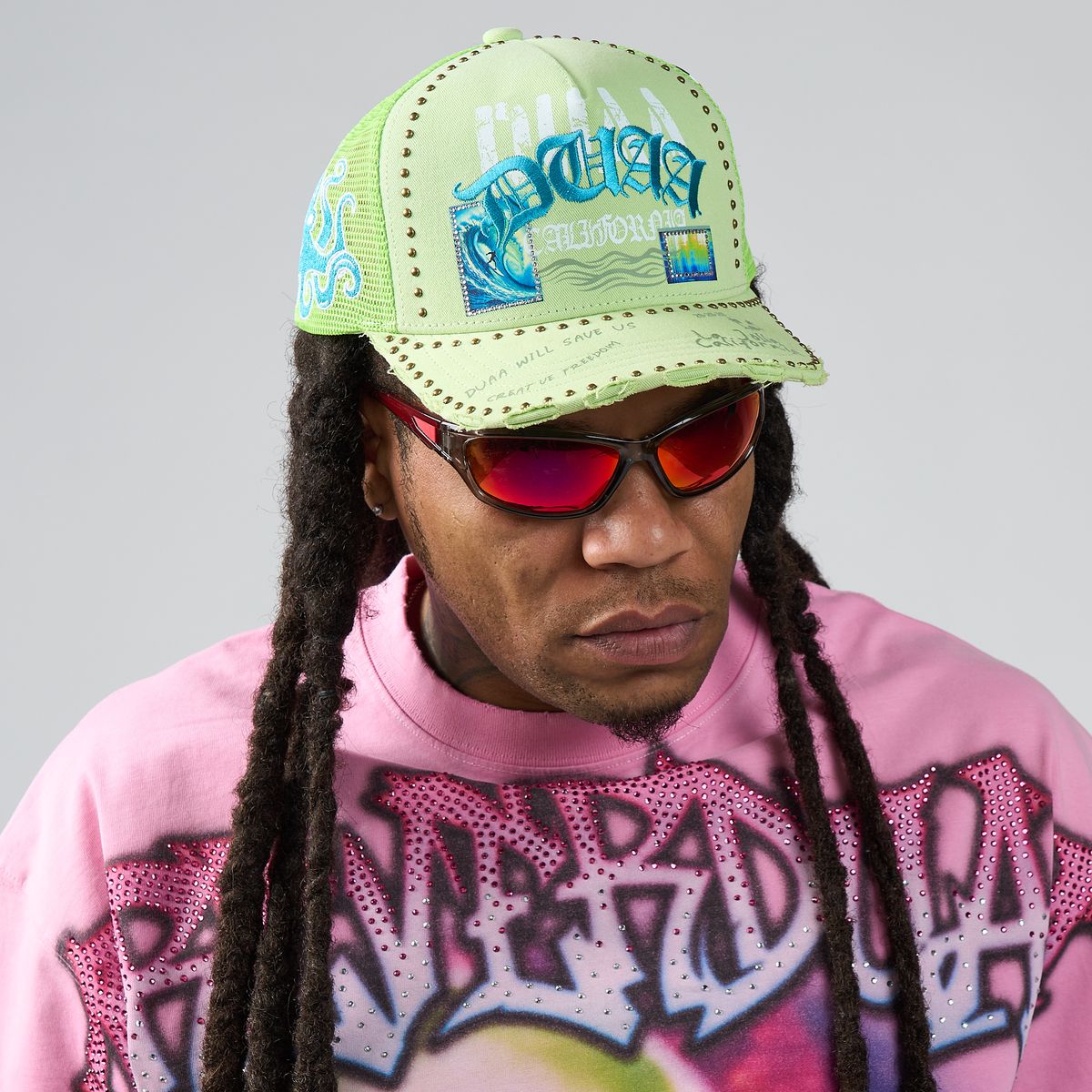 Person wearing a bright green cap with text and graphics, red sunglasses, and a pink sweatshirt with graphic designs.