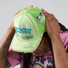 Person holding a green cap with various logos and text against a neutral background