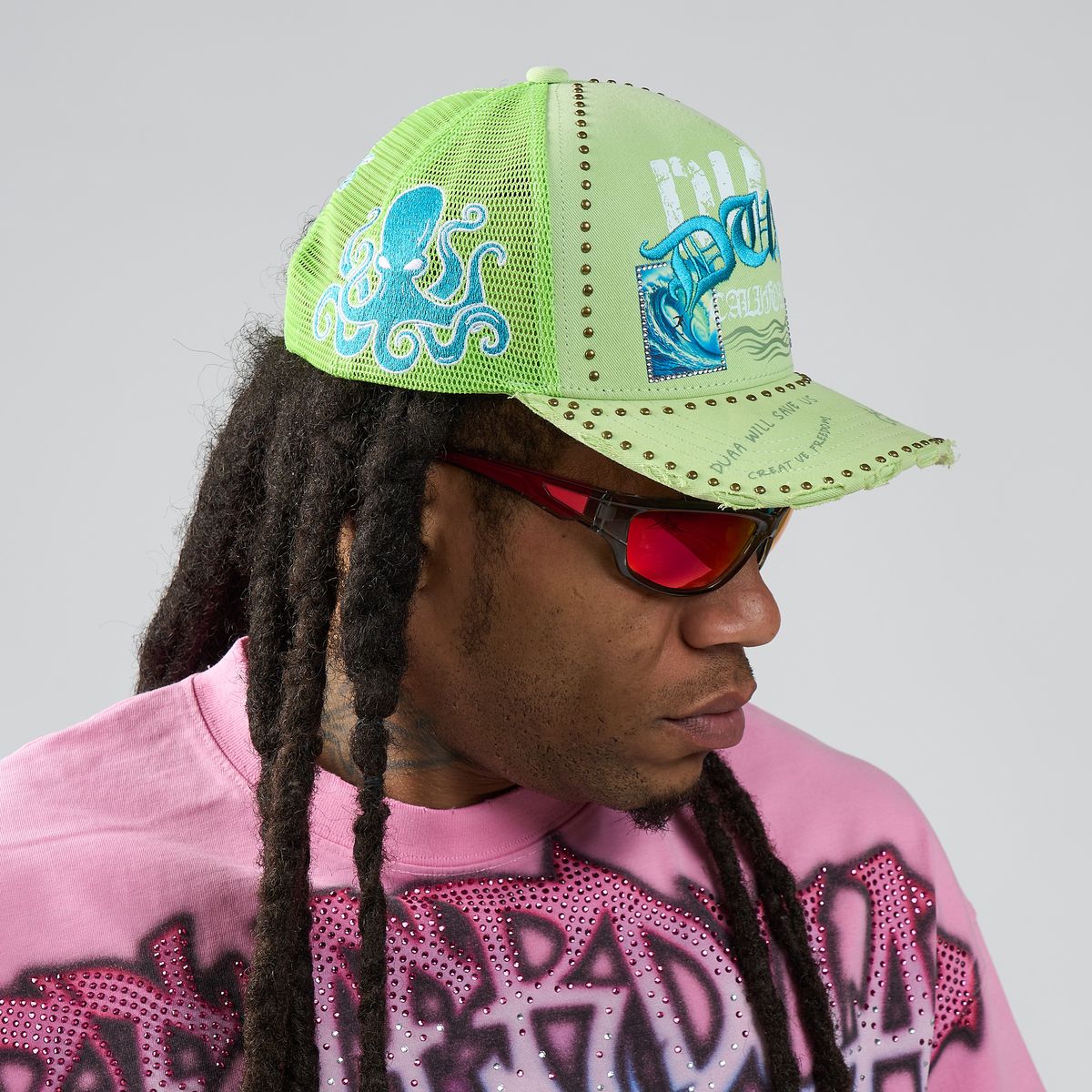 Person wearing a green cap with blue designs and red sunglasses, against a gray background