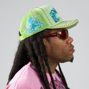 Person wearing a green cap with blue designs and sunglasses, on a plain background