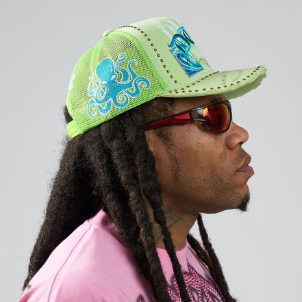 Person wearing a green cap with blue designs and sunglasses, on a plain background