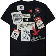 A black short-sleeve t-shirt with a collage of vintage-style graphic patches and text prints on the front, including 'Welcome to Almost Someday', 'Adventure Live Somewhere Outward', and a red star with 'Must'.