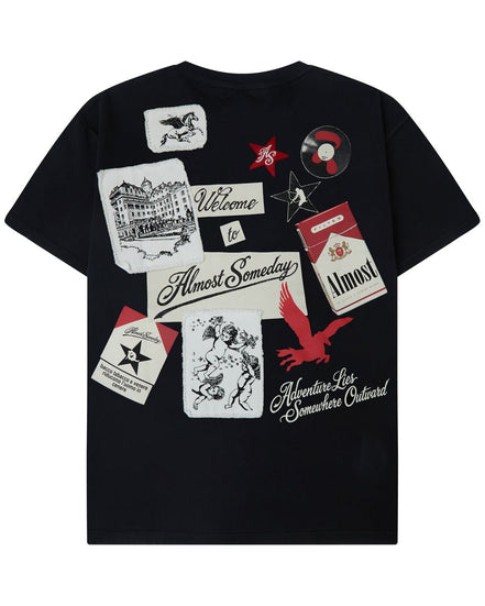 A black short-sleeve t-shirt with a collage of vintage-style graphic patches and text prints on the front, including 'Welcome to Almost Someday', 'Adventure Live Somewhere Outward', and a red star with 'Must'.