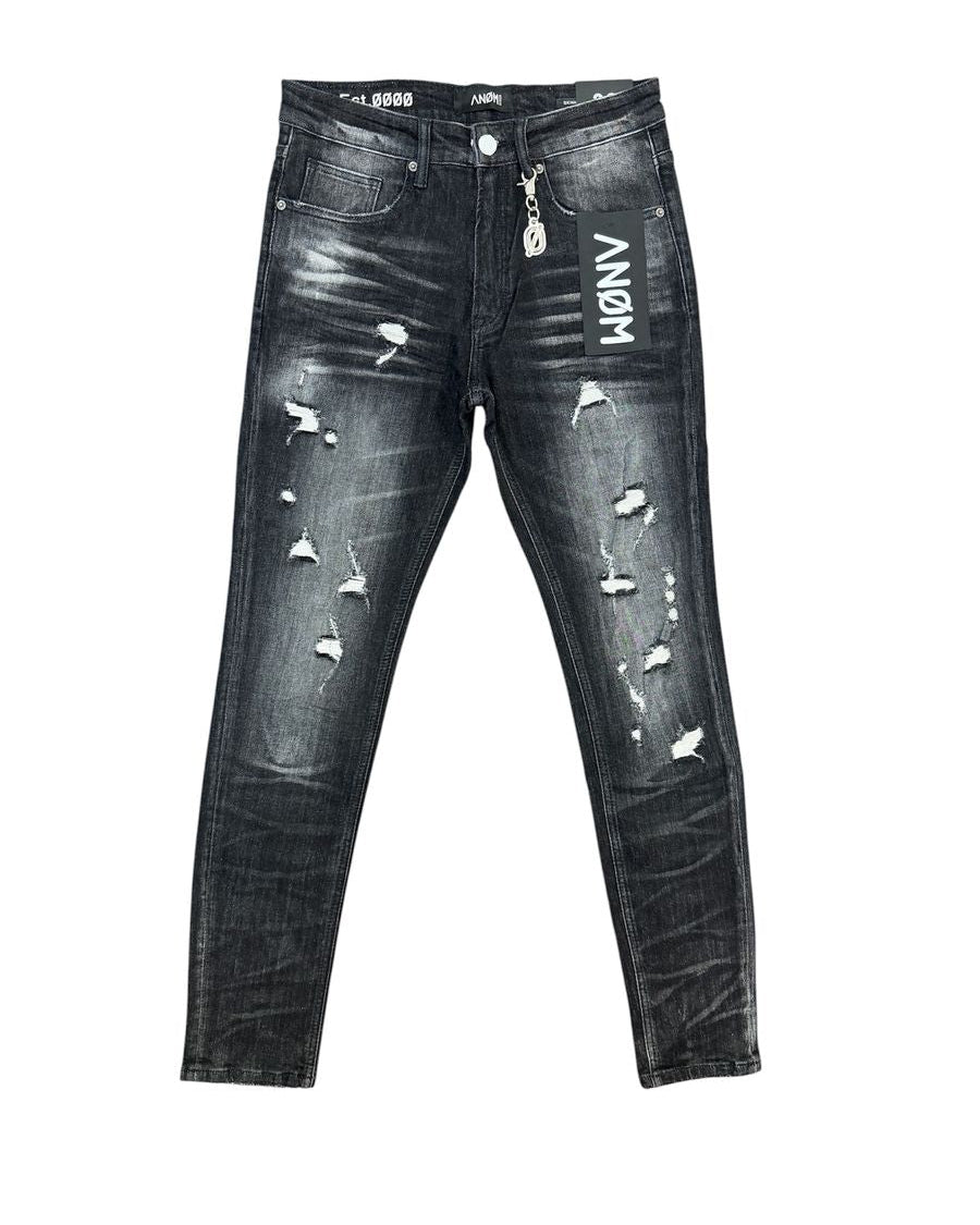 Distressed black denim jeans with white 'ANOM' lettering on the right thigh, ripped details, and a silver metal zipper pull, shown flat against a white background.