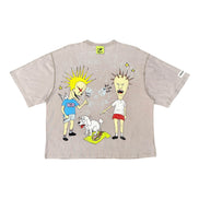 A light beige graphic t-shirt featuring a cartoon illustration of two characters and a small white dog, displayed flat against a white background.
