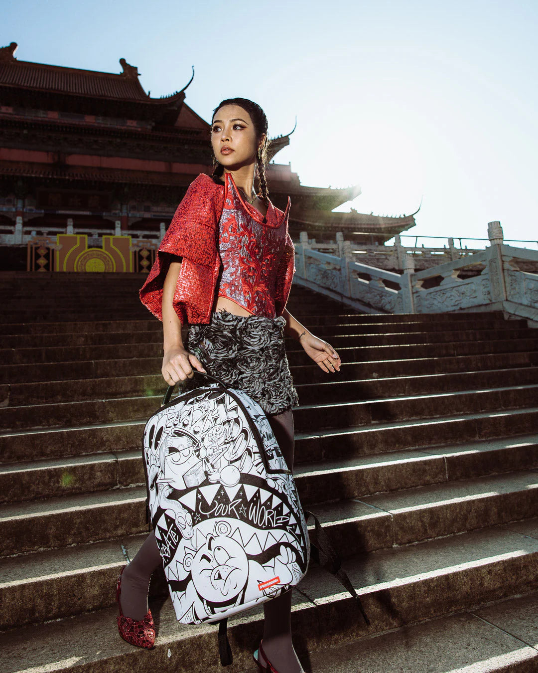 Person holding a graffiti-style Sprayground backpack on stone steps with traditional architecture in the background