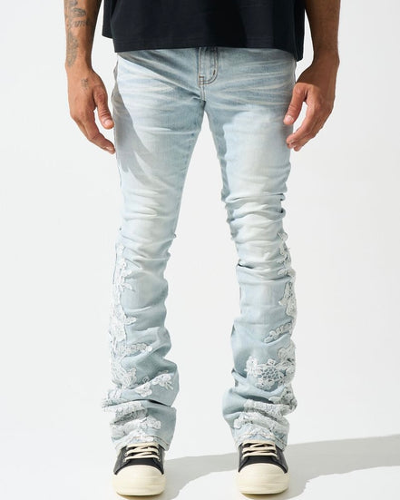 A pair of light-wash, stacked jeans featuring intricate floral lace appliques along the legs.