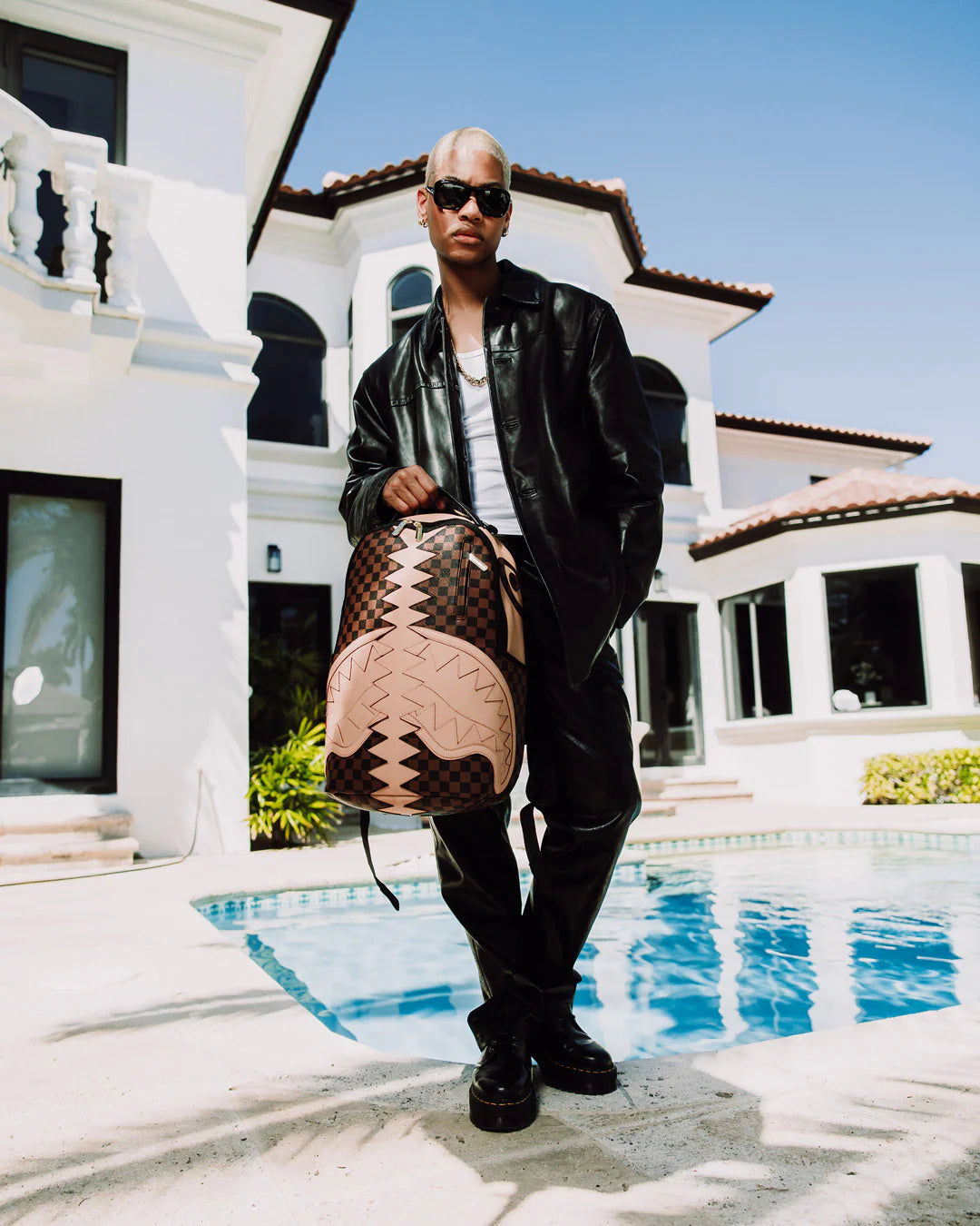 Person holding a Louis Vuitton-inspired bag, created by Sprayground, by a pool with a house in the background