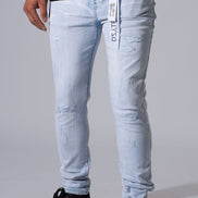 Light wash blue distressed skinny jeans with ripped details and a white vertical brand tag on the pocket, shown on a model wearing black sneakers against a gray studio background.