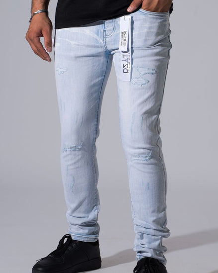 Light wash blue distressed skinny jeans with ripped details and a white vertical brand tag on the pocket, shown on a model wearing black sneakers against a gray studio background.