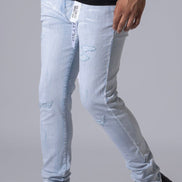 Light wash distressed denim jeans worn by a model, paired with a black t-shirt and black sneakers, shown from the waist down against a neutral gray background.