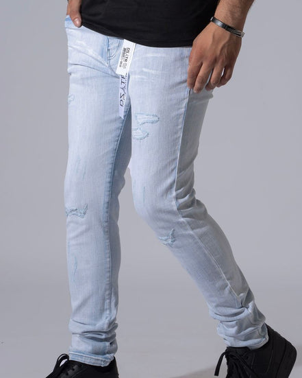 Light wash distressed denim jeans worn by a model, paired with a black t-shirt and black sneakers, shown from the waist down against a neutral gray background.
