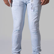 Light-wash distressed skinny jeans with a visible brand tag on the pocket, modeled with black sneakers against a neutral gray background.