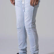 Light wash blue slim-fit jeans with distressed details and a white vertical brand tag on the pocket, worn by a model with black sneakers against a light gray background.