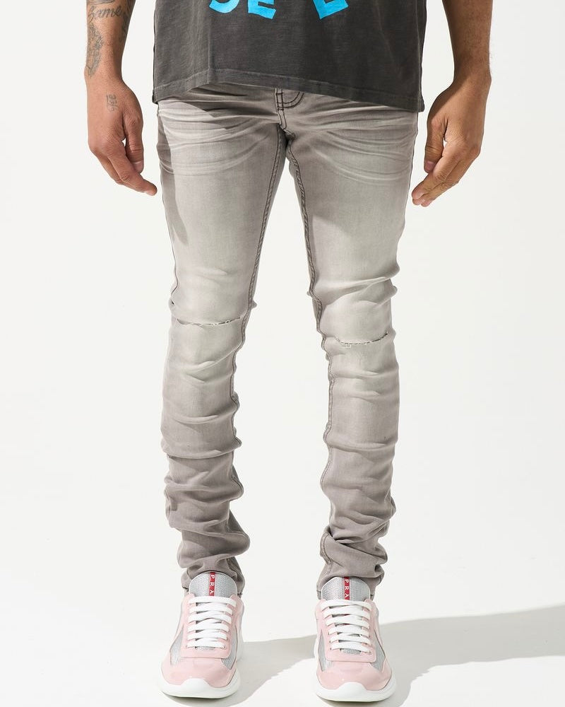 A person standing against a white background, wearing gray skinny jeans with subtle distressing on the knees and white sneakers.