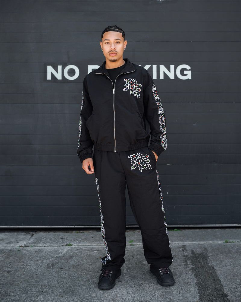 Person wearing a black tracksuit with white text against a black wall with 'NO PARKING' sign.